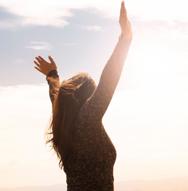 A woman with raised arms enjoying the bright open sky in a serene outdoor setting.